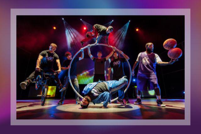 image of 4 males holding a large hula hoop type ring, 1 male in a break dancing pose in front, 1 male on a BMX bike, and 1 male juggling basketballs