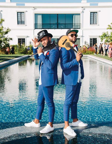 Image of two males wearing blue suits, and white shoes standing back to back in front of a swimming pool. The man on the left is holding a violin over his left shoulder and the man on the right is holding a ukulele over his right shoulder. 