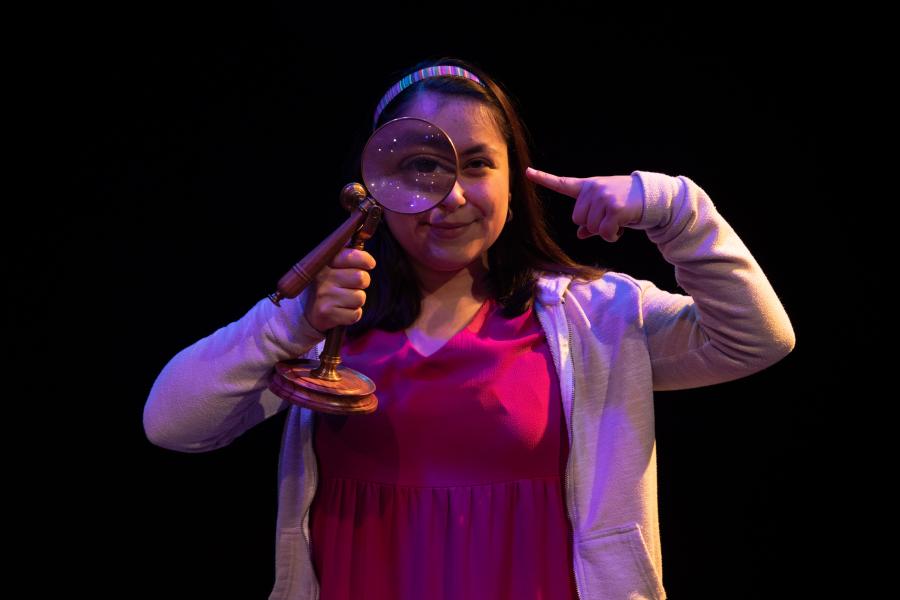 A young woman stands on stage looking through a magnifying glass while pointing to it.
