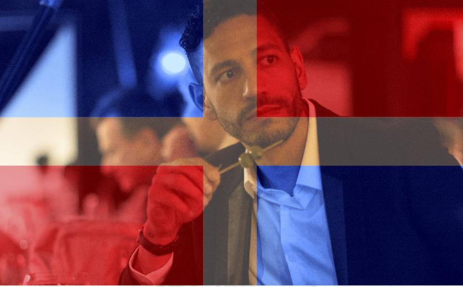 Male actor holding a martini against a red and blue flag overlaid on the image
