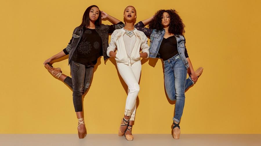 Three dancers wearing jeans and pointe shoes against a yellow background